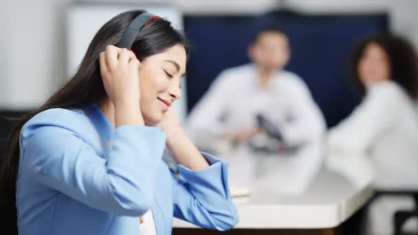 Vista lateral da bela mulher asiática despreocupada em fones de ouvido dançando ignorando colegas no escritório. Retrato de sorrir feliz empregado relaxado desfrutando de música no local de trabalho dentro de casa. — Vídeo de Stock