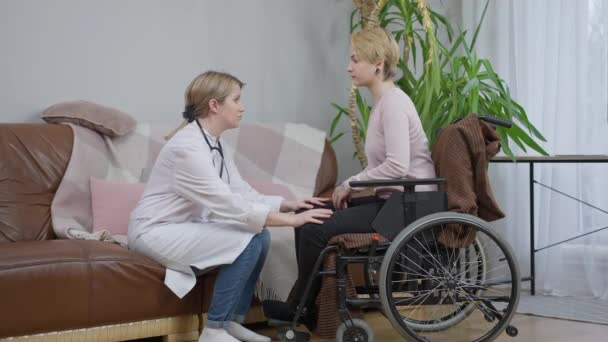 Широкий огляд професійного лікаря, який потішає інваліда. Жіночий фахівець з кавказу розмовляє з інвалідом у кріслі на колесах і торкається паралізованих ніг у повільному русі.. — стокове відео