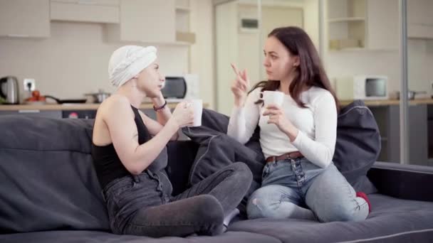 Two young beautiful positive women sitting on couch talking and drinking tea or coffee. Portrait of happy relaxed Caucasian friends resting indoors gossiping and smiling. Friendship concept. — Stock Video