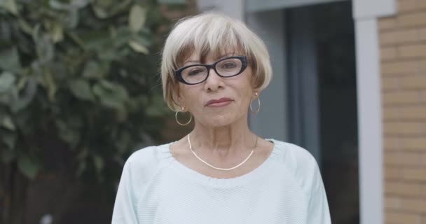 Close-up portret van stijlvolle zelfverzekerde blanke senior vrouw met bruine ogen kijkend naar de camera en glimlachend. Gelukkige gepensioneerde in een bril die buiten poseert op zomerdag. Cinema 4k ProRes Hoofdkwartier. — Stockvideo