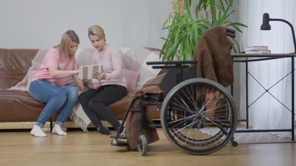 Amies positives détendues assis sur le canapé en utilisant les médias sociaux sur tablette. Vue d'ensemble de la jeune personne handicapée caucasienne et ami surfer sur Internet en ligne à la maison. Fauteuil roulant devant. Mouvement lent — Video