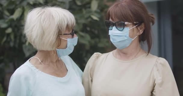 Close-up dari wanita senior Kaukasia ceria berkacamata mata dan masker wajah coronavirus melihat ke kamera. Potret dua pensiunan yang berpose di luar ruangan pada pandemi Covid-19. Sinema 4k ProRes HQ. — Stok Video