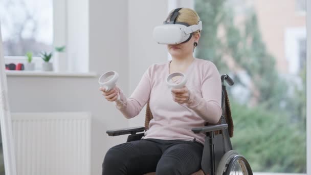 車椅子の室内に仮想現実ヘッドセットを使用して積極的な若い超常現象女性。VRヘッドセットとモーションコントローラーを使用して肯定的な白人障害者の肖像画オンラインゲーム. — ストック動画
