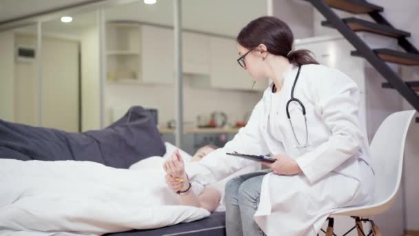 Brunette Caucasian vrouw in dokters jurk en bril nemen pols van uitgeputte jonge vrouwelijke patiënt liggend op de bank thuis. Professionele ambulance arts die zorgt voor vrouw met kanker. — Stockvideo