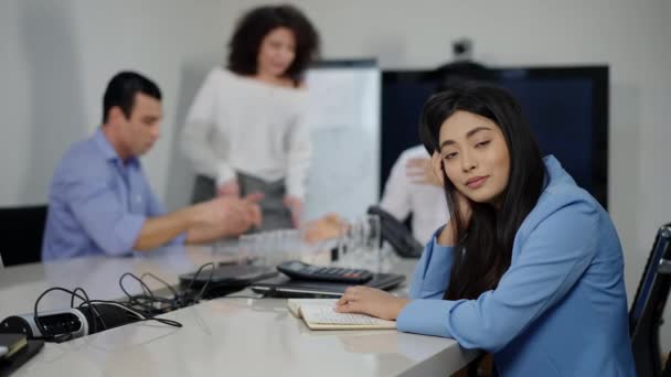 Encantadora mulher de negócios asiática magra sentada na mesa de conferência balançando a cabeça como colegas desfocados discutindo no fundo. Jovem estressada posando no cargo durante brainstorming. Conceito de trabalho em equipa. — Vídeo de Stock