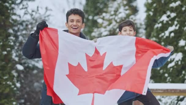 Gelukkige opgewonden Midden-Oosterse tiener en jongen met Canadese vlag die buiten staan op besneeuwde dag. Portret van vrolijke broers die poseren met nationaal symbool in Canada. Immigratie en levensstijl. — Stockvideo