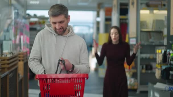 Portret zadowolonego białego mężczyzny patrzącego w kamerę i uśmiechającego się jako podrażniona kobieta krzycząca i gestykulująca w tle. szczęśliwy mężczyzna shopper pozowanie w sprzęt sklep z irytujące żona krzyczy. — Wideo stockowe