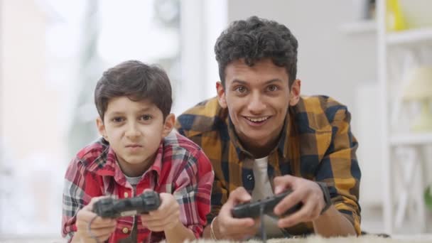 Ontspannen Midden-Oosten broers gaming met behulp van game controllers als sluwe kind sluiten ogen van tiener om te winnen. Lastige jongen vals spelen online met broer of zus. Vrije tijd en ontspanning. — Stockvideo