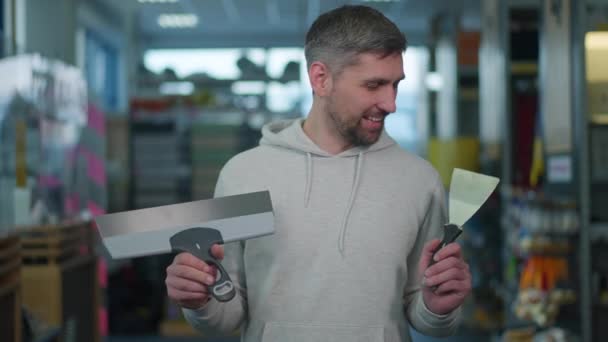 Hombre caucásico barbudo eligiendo cuchillo masilla en ferretería. Retrato del comprador masculino guapo que toma la decisión en la tienda de suministros de construcción. Elección y concepto de renovación. — Vídeos de Stock