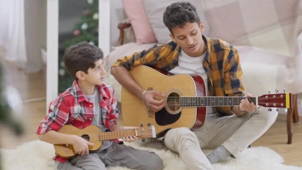 Positiva bröder från Mellanöstern som spelar gitarr och ukulele i slow motion-sång. Bred bild av leende tonåring och barn öva musik hemma tillsammans. Konst och livsstil. — Stockvideo