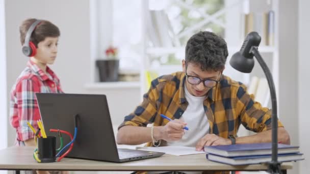 Ocupado adolescente concentrado en anteojos ignorando a su hermano pequeño distrayendo al estudiante de la tarea. Molesto adolescente de Oriente Medio sentado en la mesa estudiando como triste hermano dejando. — Vídeos de Stock