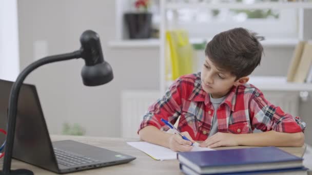 Cute focused Middle Eastern boy writing homework and shaking head yes in video chat on laptop. Portrait of concentrated obedient schoolboy studying remotely on pandemic lockdown. Slow motion. — Stock Video