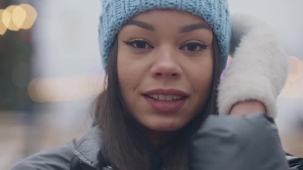 Cara de cerca de una joven afroamericana sonriente con sombrero azul de invierno y manoplas blancas posando en un día nevado de invierno al aire libre. Cabeza de positivo feliz relajado millennial disfrutando de clima frío. — Vídeo de stock