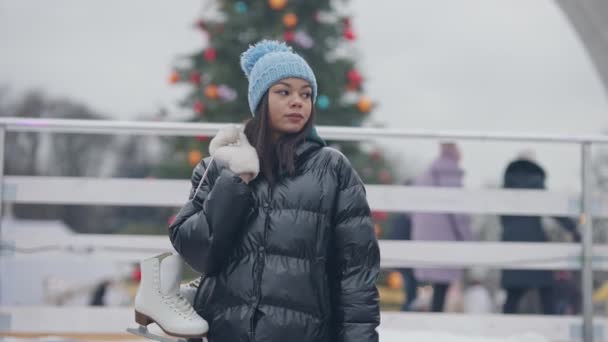 Porträtt av självsäkra tusenåriga hängande skridskor på axeln tittar runt på snöiga vinterdag. ung vacker afroamerikansk kvinna vilar utomhus med suddig ishall i bakgrunden. — Stockvideo