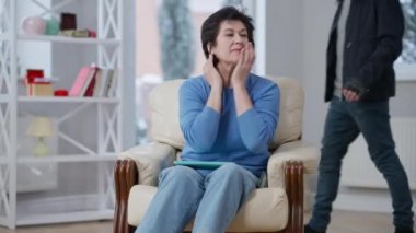 Endişeli soygun kurbanının portresi, arka planda yürüyen maskeli bir hırsızla koltukta oturuyor ve silahla tehdit ediyor. Korkak Kafkasyalı kadın evde silahlı bir hırsız tarafından soyuldu.