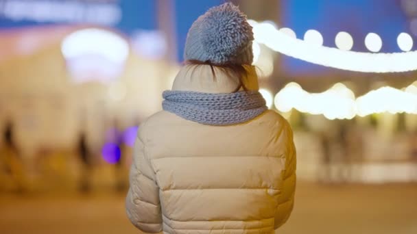 Widok z tyłu szczęśliwej beztroskiej młodej kobiety stojącej w nocnym mieście podziwiającej lampki świąteczne. Bliski strzał zrelaksowanego Kaukaskiego tysiąclecia cieszącego się wieczornym miejskim wypoczynkiem na zewnątrz. — Wideo stockowe