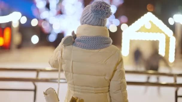 Widok z tyłu wesołej młodej kobiety z łyżwami na ramieniu obracającej się do kamery ze śmiechem. Portret radosnego białego tysiąclecia odpoczywającego na nocnym zimowym miejskim lodowisku na świeżym powietrzu. — Wideo stockowe