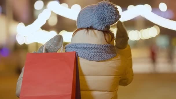 Baksidan av avslappnad vit ung shoppare står på vintern natt utomhus med shoppingväskor hängande på axeln. Obekymmerslös shopaholic på stadskvällen. Livsstil och shopaholism. — Stockvideo