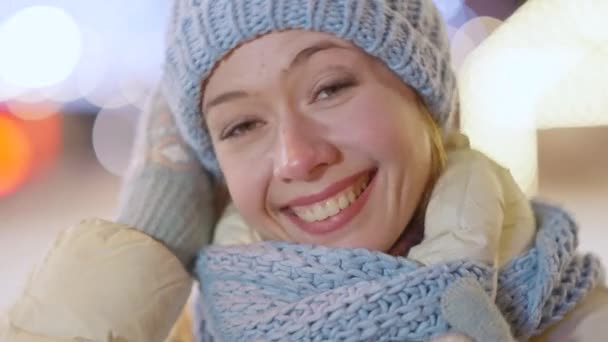 Zdjęcie uśmiechniętej młodej pięknej kobiety z brązowymi oczami patrzącej na kamerę stojącą na zewnątrz w zimową noc. Close-up portret szczęśliwy zrelaksowany biały tysiąclecia z miejskich świateł w tle. — Wideo stockowe