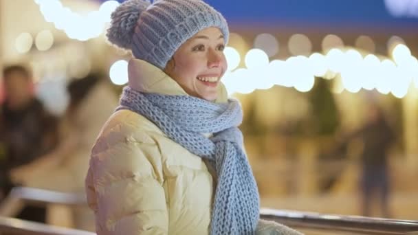 Zijaanzicht van vrolijke jonge vrouw met tanden glimlach zwaaiend en wegkijkend staande op de winter ijsbaan in de stad. Portret van gelukkige positieve Kaukasische duizendjarige ontmoeting vriend op stedelijke nacht buiten. — Stockvideo