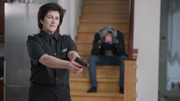 Портрет уверенной женщины-полицейского, поднимающей руки с пистолетом, смотрящей в камеру, как несчастный преступный вор сидит на лестнице на заднем плане. Кавказка арестовывает грабителя в богатом доме. — стоковое видео