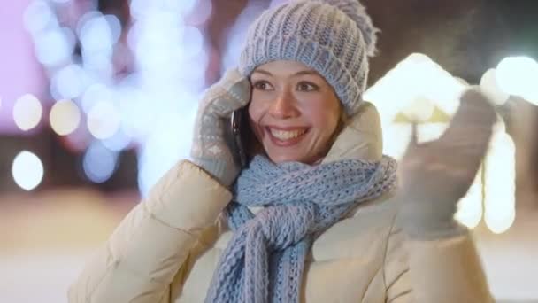 Portret van vrolijke jonge slanke blanke vrouw die aan de telefoon praat en buiten zwaait in de nachtstad. Vrolijke vrolijke millennial wachtend op vriend op stedelijke avondstraat. Levensstijl concept. — Stockvideo