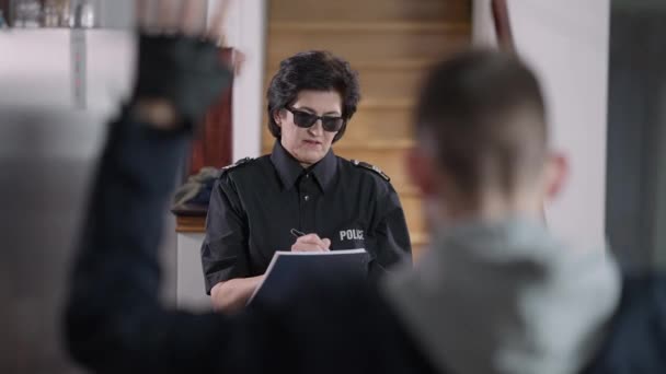 Una mujer policía sonriente con gafas de sol hablando con el borroso ladrón arrestado al frente. Retrato de una policía caucásica satisfecha atrapando a un ladrón peligroso rellenando el informe. Proteger y servir. — Vídeos de Stock