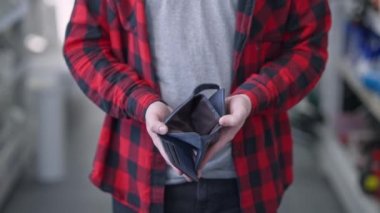 Erkek ellerinin boş cüzdanı açtığı yakın plan. Tanımlanamayan beyaz bir adam dükkânda parası olmayan, bulanık sıraların arasında duruyor. Tüketim ve yoksulluk.