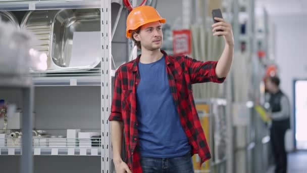 Hombre guapo alegre tomando selfie en la ferretería. Milenario morena sonriente positiva en sombrero duro tomando fotos para las redes sociales en la construcción y renovación de la tienda de suministros. Concepto de estilo de vida. — Vídeo de stock