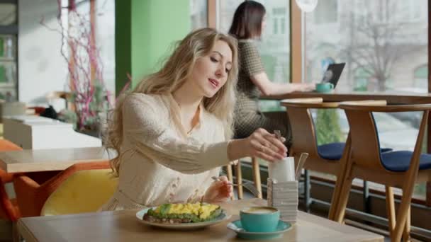 Ontspannen slanke blanke vrouw eten delicatessen in cafe met wazig zakenvrouw typen op laptop op de achtergrond. portret van gelukkig jong blond millennial rusten in restaurant op zonnige dag. — Stockvideo
