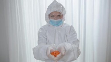 Koruyucu takım elbiseli, gözlüklü ve elleri turuncu haplarla dolu beyaz kadın portresi. İçeride ilaçla poz veren profesyonel doktor ya da hemşire..