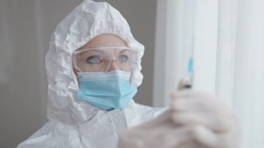 Kafkasyalı kadın doktorun yakın plan portresi şırıngayı ilaçla hazırlıyor. Koruyucu giysili, gözlüklü ve koronavirüs aşısıyla maskeli kadın..