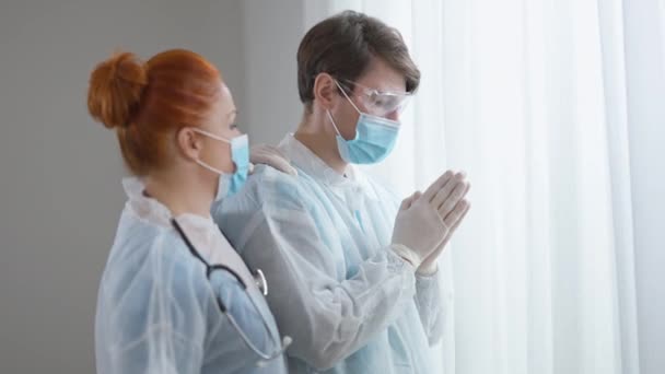 Boční pohled na empatickou sestru podporující doktora modlícího se uvnitř. Vážný běloch a žena v lékařské uniformě v nemocnici na propuknutí pandemie koronaviru. Koncept naděje a víry. — Stock video