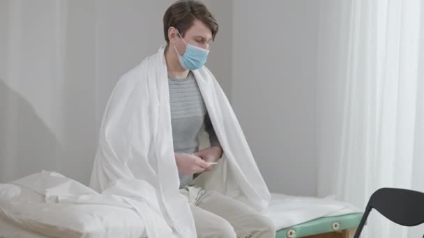 Portret van een zieke blanke man in coronavirus masker meten temperatuur met thermometer zittend op medische bank binnen. Trieste geconcentreerde mannelijke patiënt in het ziekenhuis. Gezondheidszorgconcept. — Stockvideo