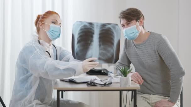 Covid-19の顔のマスクに集中した白人女性と男性は肺のX線を指して話しています。医師と患者は、コロナウイルス病や喫煙の結果を議論病院に座って.医学. — ストック動画