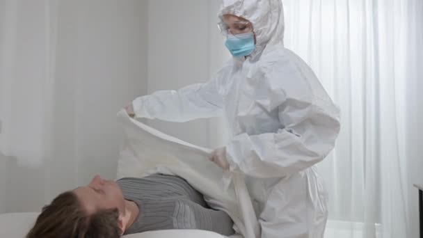 Медсестра в защитном костюме, очках и маске, покрывающей мертвого человека белым листом. Портрет грустной белой женщины-врача, потерявшей пациента от пандемии коронавируса. Концепция летального исхода. — стоковое видео
