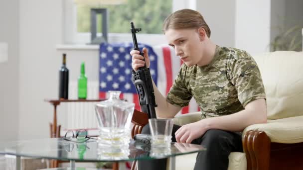 Frustrated young American soldier sitting at home holding rifle gun and looking at camera. Portrait of depressed man with weapon and national flag at background getting drunk indoors. Crisis despair. — Stock Video