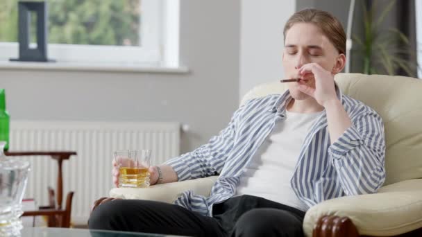 一个悠闲富有的年轻人坐在扶手椅上抽雪茄，喝苏格兰威士忌的画像。自信的白人富人在家里休息。时尚和奢侈概念. — 图库视频影像