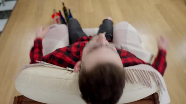 Ovanifrån av berusad ung man i glasögon lutar sig mot fåtölj med suddiga alkoholflaskor står på golvet i bakgrunden. Fylld kaukasisk alkoholist sittande på soffan inomhus. — Stockvideo