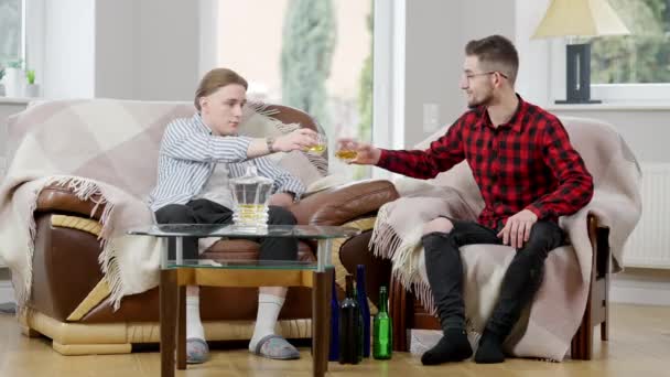 Weitwinkelaufnahme von zwei entspannten kaukasischen Männern, die Gläser klirren und Whisky trinken. Porträt männlicher Freunde, die mit Alkohol zu Hause ausruhen. Freundschaft und Alkoholismus. — Stockvideo