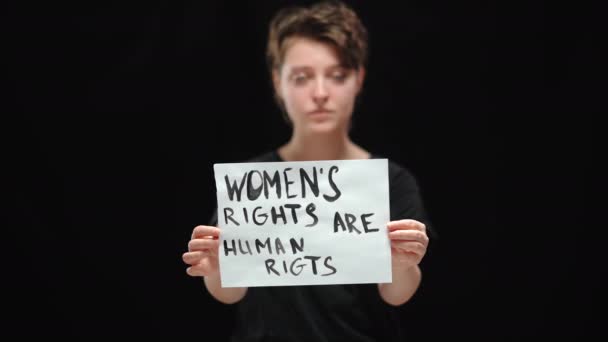 Primer plano del cartel Los derechos de las mujeres son derechos humanos con una mujer joven seria borrosa de fondo negro. Una activista confiada que lucha por la igualdad de derechos. Feminismo y feminidad. — Vídeos de Stock