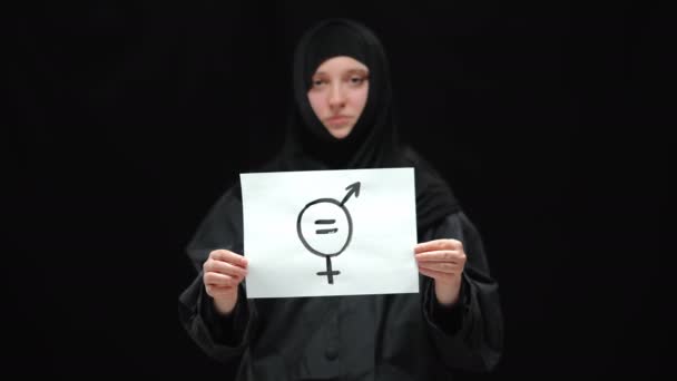 A nemek közötti egyenlőség közeli jele a háttérben a hidzsábi homályos nővel. Szomorú, fiatal muszlim nő, fekete háttérben pózol. Nőiesség és egyenlő emberi jogi koncepció. — Stock videók