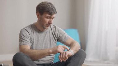 Tatmin olmuş gülümseyen adamın portresi spor salonundaki eğitim sonuçlarını kontrol ediyor. Kafkasyalı mutlu orta yaşlı sporcu akıllı saatini evde otururken kullanıyor. Antrenman ve spor.