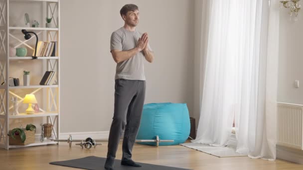 Weitwinkelporträt eines ruhigen, selbstbewussten Sportlers, der in Baumpose auf einer Trainingsmatte im Innenraum steht. Kaukasischer Mann mittleren Alters trainiert morgens Yoga zu Hause. Yogi-Training. — Stockvideo