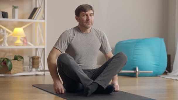 Portret spokojnego mężczyzny w średnim wieku w lotosie stanowi medytację w pomieszczeniach. Przystojny, pewny siebie, biały sportowiec ćwiczący jogę w domu. Medytacja i spokój. — Wideo stockowe