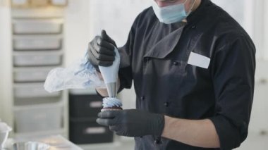 Covid-19 yüz maskesi takmış, fırında pişmiş kekin üzerine mavi tatlı döken tanınmamış bir beyaz adam. Şekerci dükkanında pasta süsleyen profesyonel bir erkek şekerci..