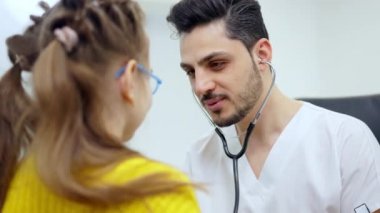 Steteskop kullanan ve konuşan güzel, pozitif çocuk doktorunun portresi. Neşeli Orta Doğulu adam hastanedeki küçük beyaz hastaya danışıyor. Pediyatri Kliniği.