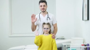 Pozitif Orta Doğulu erkek çocuk doktoru portresi ve kameraya el sallayan gözlüklü sevimli beyaz kız. Hasta ve doktor hastane koğuşunda poz veriyor. Tıbbi konsept.