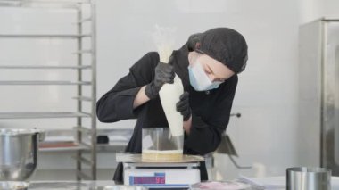 Mutfak terazisinde duran fırında kek hamuruna katman ekleyen odaklanmış kadın şekerlemecinin portresi. Coronavirus yüzlü genç beyaz kadın içeride tatlı pişiriyor..