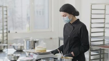 Genç, sıska kadın pastacı. Önünde ticari tepsisi olan bulanık bir adam olarak fırında kek katmanı ekliyor. Covid maskeli, tatlı pişiren beyaz kadın portresi..